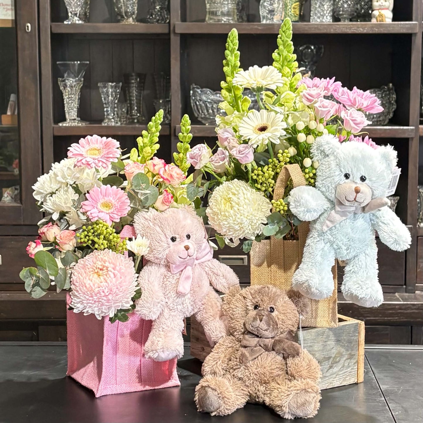Three plush teddy bears in pink, brown, and light blue are arranged with colorful flower bouquets in pink, wood, and gray fabric baskets in front of a dark wooden display cabinet.