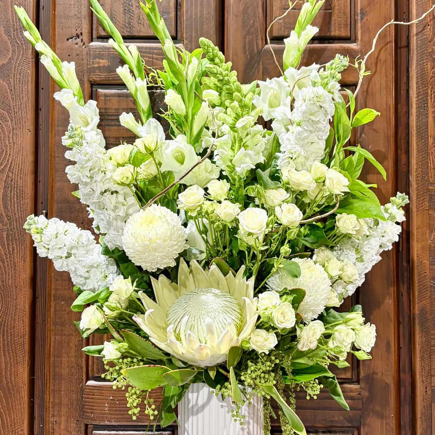 
                  
                    A large floral arrangement featuring white protea, hydrangea, roses, snapdragons, and green foliage in a white ribbed vase, set against a dark brown wooden door background.
                  
                