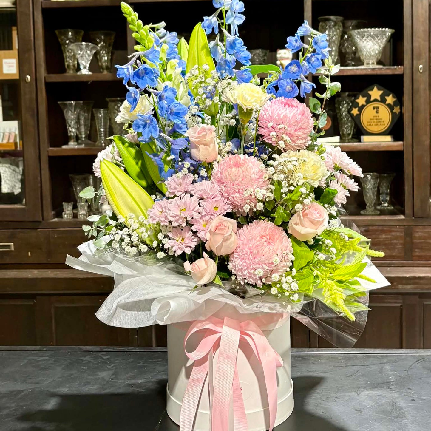 
                  
                    Bouquet of flowers in a white container with pink ribbon on a dark surface, with a wooden cabinet in the background.
                  
                