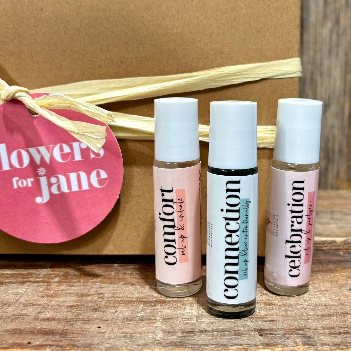 Three rollerball bottles labeled 'Comfort', 'Connection', and 'Celebration' in front of a cardboard box with a 'flowers for jane' tag.