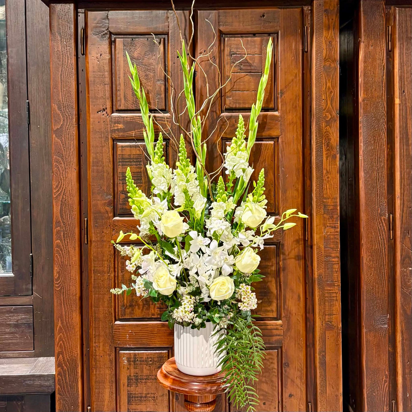 A large floral arrangement featuring white and pale yellow flowers with green foliage, displayed in a white ribbed ceramic pot on a wooden pedestal in front of a dark wood paneled door.