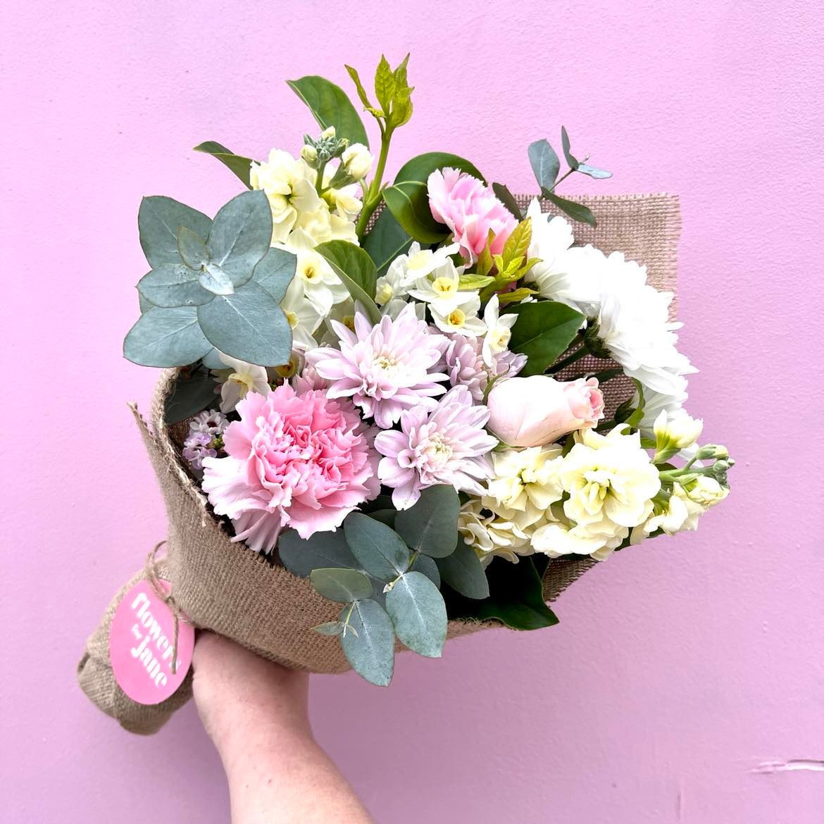 A bouquet of flowers wrapped in white paper, with a mix of pink, white, and green flowers, held against a pink background.