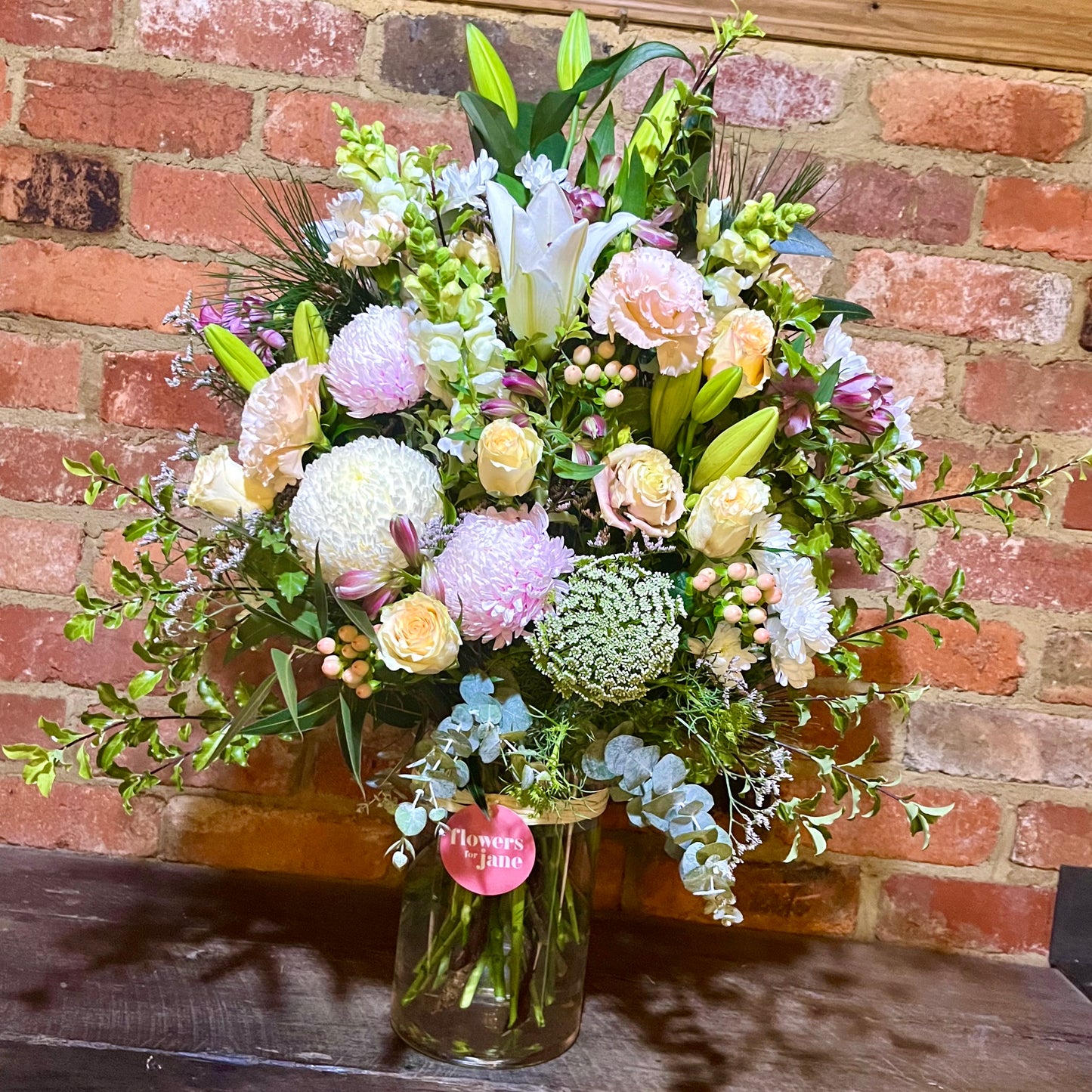 A vase of fresh-cut seasonal blooms with a variety of colors including red, pink, white, and green, arranged in a glass vase placed on a wooden surface.